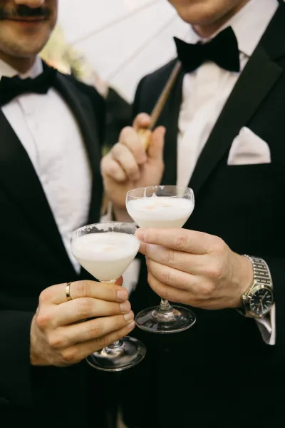 Cocktail hour cheers at the wedding cocktail hour as coupe glasses clink beneath an umbrella, lush greenery framing the celebratory toast.