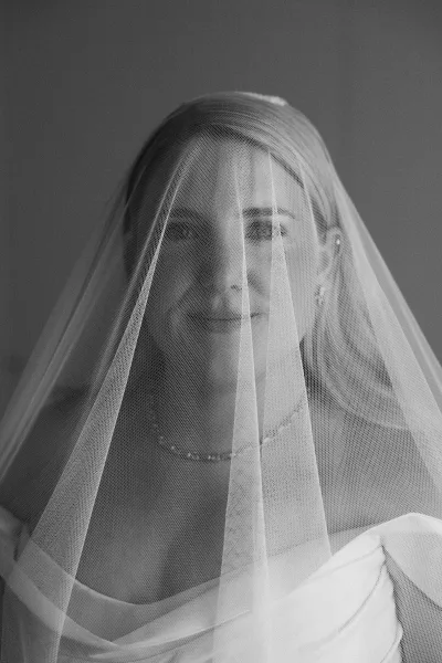 Black and white bridal portrait of a bride under veil, revealing an off-shoulder gown as she gazes softly with timeless poise.