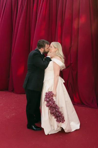 Wedding kiss portrait of a couple embracing, bride holding an orchid bouquet against rich red draped fabric and a matching red carpet.