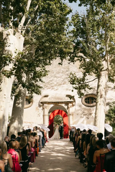 Ceremony moment as the couple shares vows during a courtyard wedding ceremony, framed by trees and a stucco wall, beneath a red drape.
