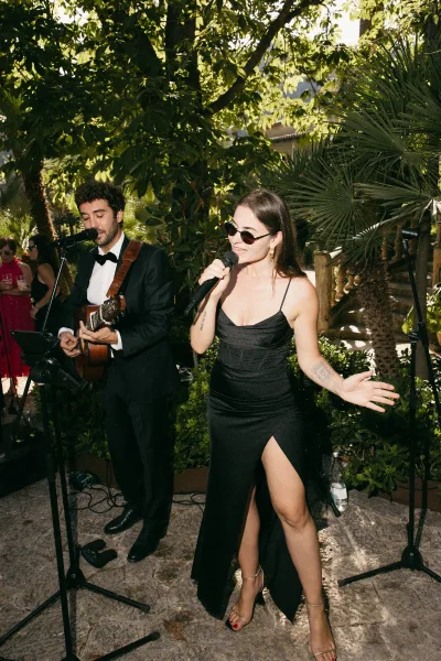 Live wedding music duo plays cocktail hour music, singer at a microphone beside a guitarist on a stone patio framed by garden trees and palm leaves.
