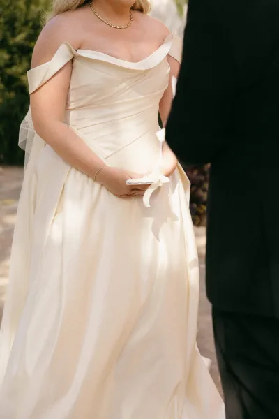 Ceremony moment during a wedding vows exchange as the bride reads from a ribbon-tied vow book beside the groom, in dappled garden greenery.