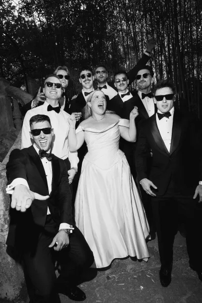 Wedding party portrait of a bride with groomsmen wearing sunglasses, gathered on stone steps within a bamboo grove and lush garden foliage.
