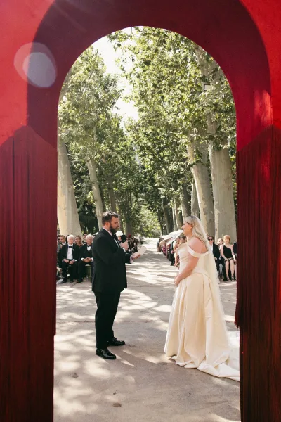 Ceremony moment during an outdoor wedding ceremony as the bride and groom face each other beneath a bold red arch along a sunlit tree-lined path.