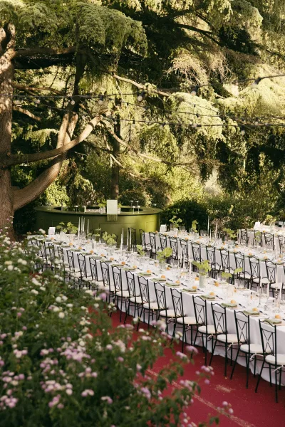 Reception tablescape for an outdoor garden reception, long banquet tables beneath garden trees accented with taper candles for a warm, elegant glow.