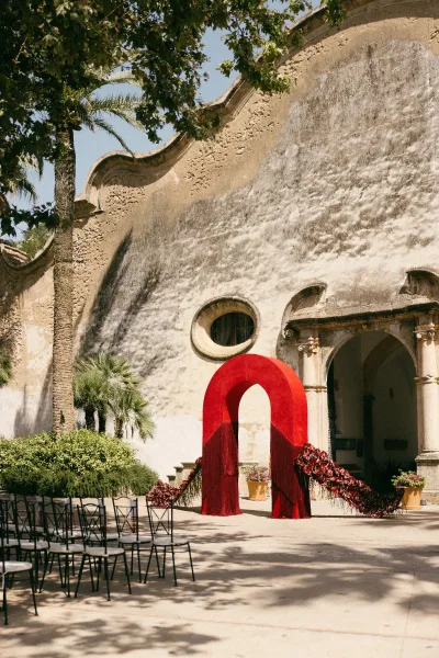 Ceremony arch anchors the outdoor ceremony decor with bold red florals before a stucco arched doorway in a palm-lined courtyard.