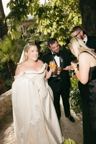 Wedding cocktail hour with a bride holding a cocktail laughing with her groom along a stone path beneath palm trees and villa arches.