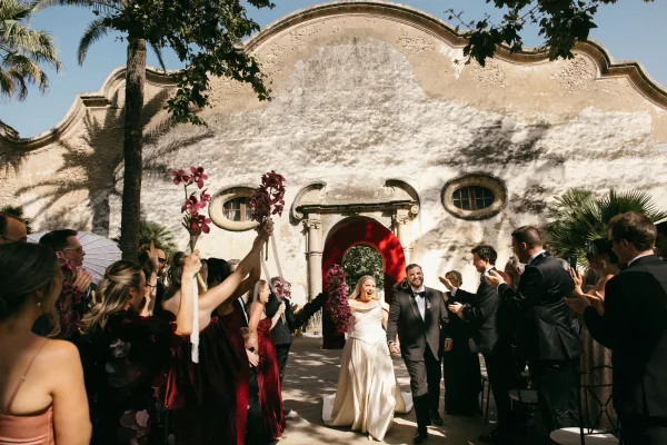 Ceremony recessional as the couple holding hands walk beneath a stucco arch and palm trees, the bride lifting an orchid bouquet.