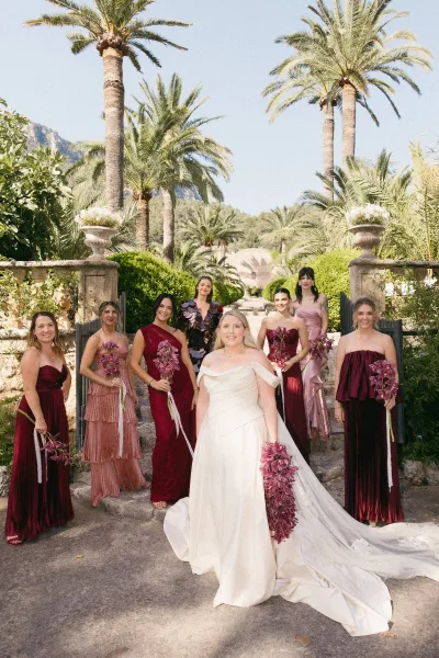 Bride with bridesmaids in burgundy bridesmaid dresses hold orchid bouquets on stone steps beneath palm trees in a lush garden setting.