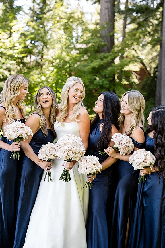 Bride with bridesmaids in navy dresses holding blush rose bouquets, gathered in sunlight among trees and greenery outdoors