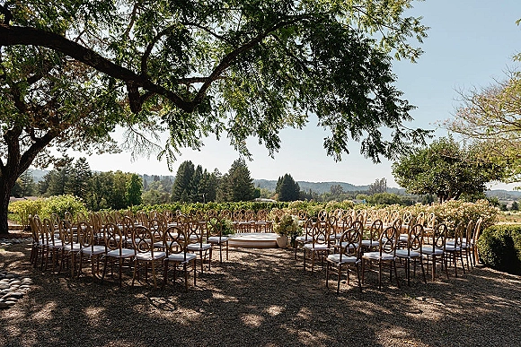 Outdoor ceremony setup with garden wedding ceremony seating in a semicircle of wood chairs around a low platform under a tree canopy in a vineyard