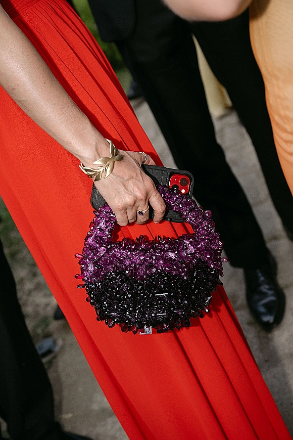 Wedding guest accessories with a beaded purple handbag and smartphone held against a red pleated dress on an outdoor walkway with guests