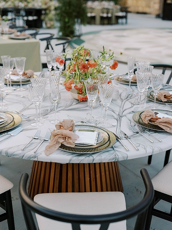 Reception tablescape with wedding place setting, gold chargers and crystal stemware, floral centerpiece on a patterned cloth on an outdoor patio table