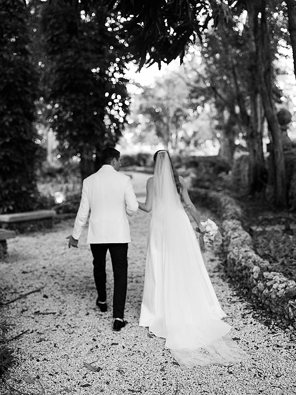 Couple portrait of bride and groom walking away holding hands, her long veil and bouquet trailing on a tree-lined garden path