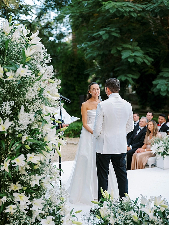 Wedding vows at an outdoor wedding ceremony as bride in a strapless dress and veil holds hands with groom in white jacket under a lily arch