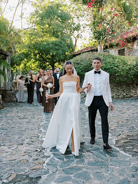 Newlywed exit as couple walking hand in hand, holding champagne flutes, with bridesmaids behind on a stone walkway in a garden courtyard