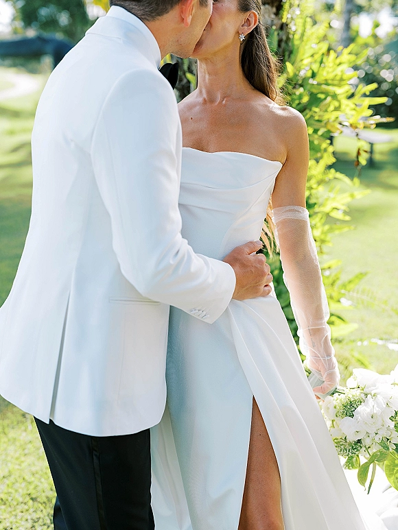 Wedding kiss portrait of bride and groom kissing, her strapless slit gown and sheer gloves, his white tuxedo, in a garden setting