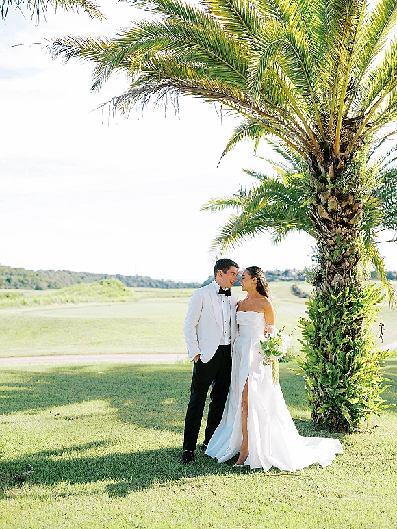 Couple portrait of bride in a strapless slit gown holding a bouquet beside groom in a white dinner jacket under a palm tree lawn and hills