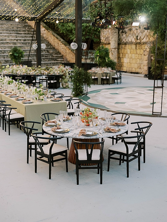 Reception tablescape with outdoor reception tables, mixed round and long settings with florals, candle votives, and bistro lights by a stone wall