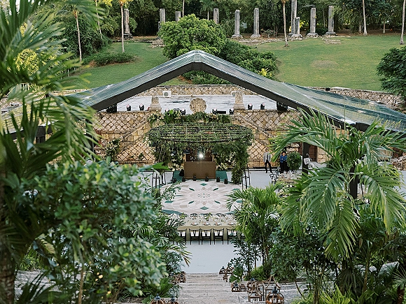 Outdoor reception setup with clear top tent reception, string lights, long banquet tables and dance floor set among palms and stone walls