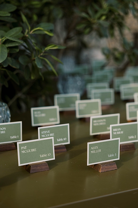 Wedding place cards in wooden holders with printed names on a glass tabletop, styled with green place cards and lush greenery backdrop