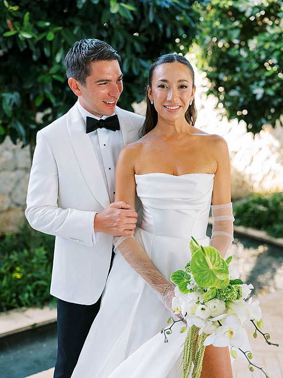 Couple portrait of bride in strapless wedding dress holding anthurium and orchid bouquet beside groom in white tuxedo in garden greenery