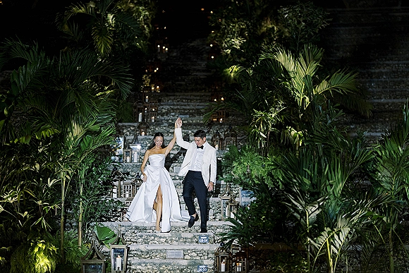 Couple portrait of bride in a strapless wedding dress and groom in white tuxedo holding hands on a candlelit stone staircase at night