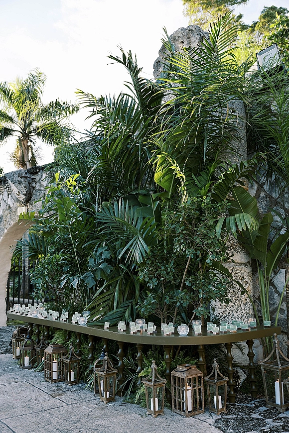 Escort card display with wedding escort cards on a long table, framed by lanterns and pillar candles against a stone wall patio backdrop