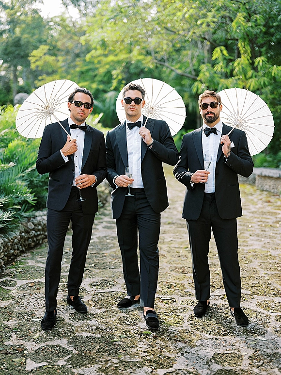 Groomsmen portrait of three men in black tuxedos and sunglasses holding white parasols on a cobblestone garden path near a stone wall
