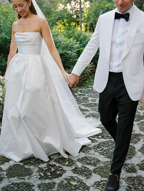 Couple portrait of bride and groom walking hand in hand, her veil flowing, on a stone pathway through lush garden greenery