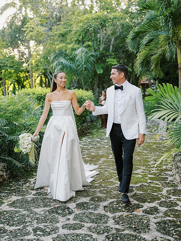 Couple portrait of bride and groom walking hand in hand, bride holding a bouquet, along a stone path in a tropical garden with palms