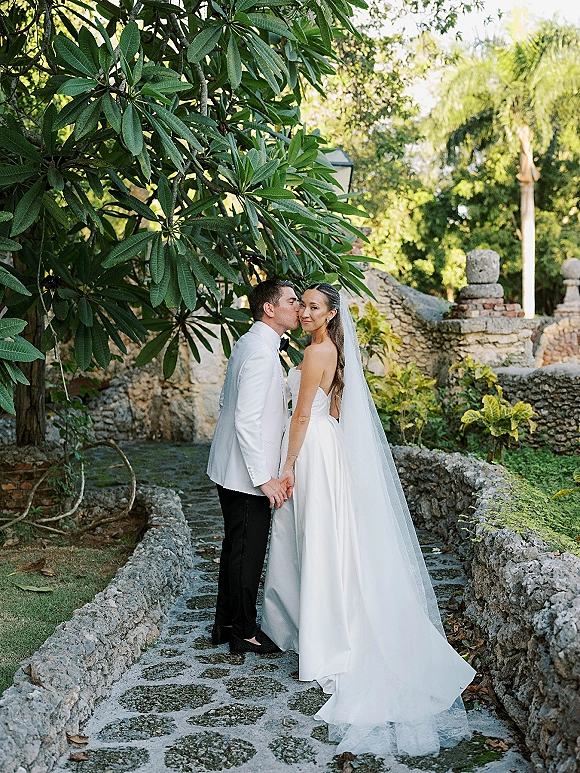 Couple portrait of bride and groom holding hands as he kisses her cheek, long veil and tuxedo on a stone garden path