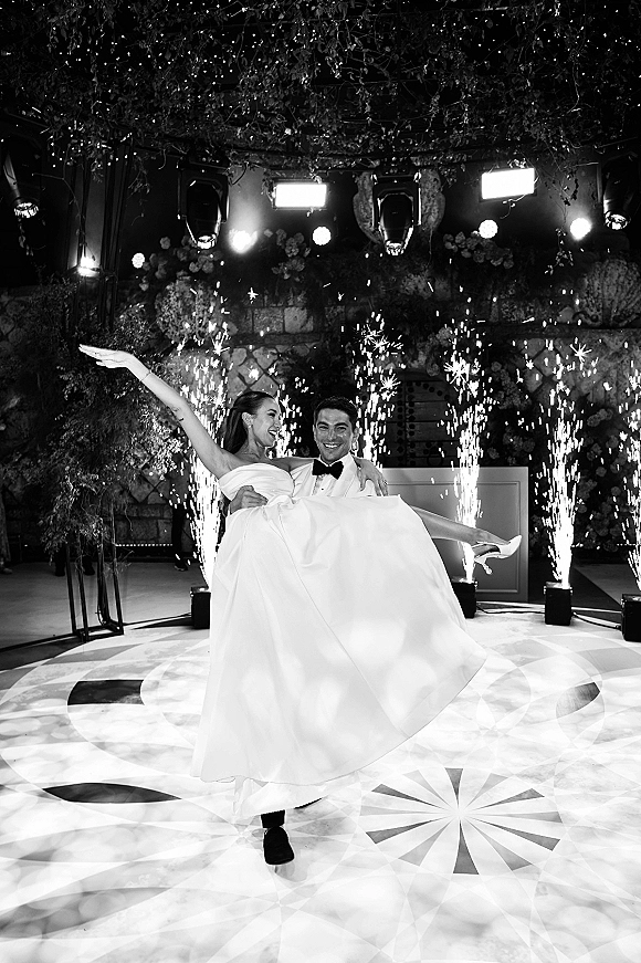 Wedding reception entrance as groom carries bride in strapless gown through sparkler fountains on dance floor under string lights and greenery