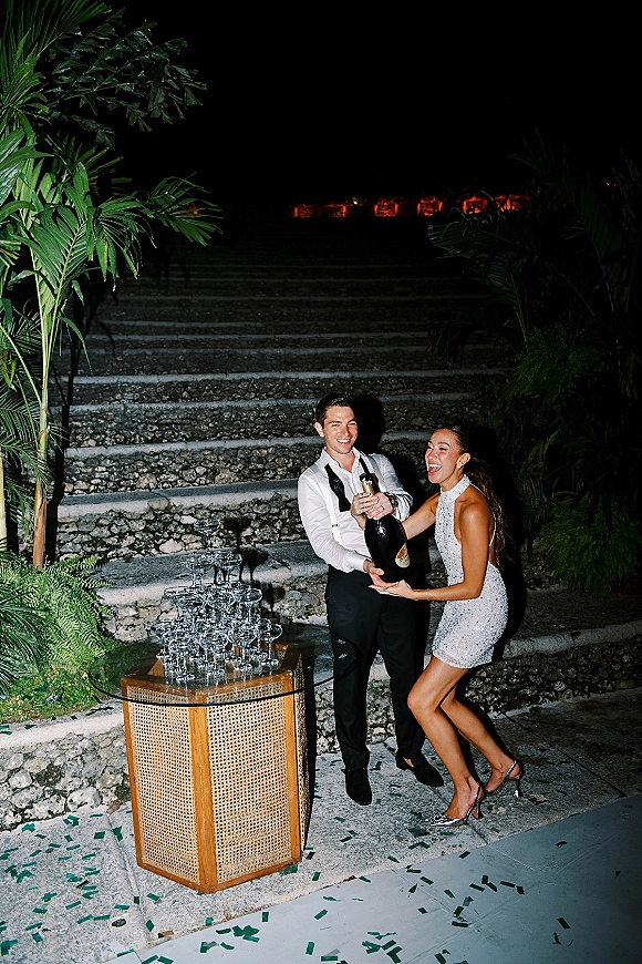 Champagne celebration at a wedding champagne pop with couple by a coupe-glass tower on stone steps under tropical greenery at night, confetti falling