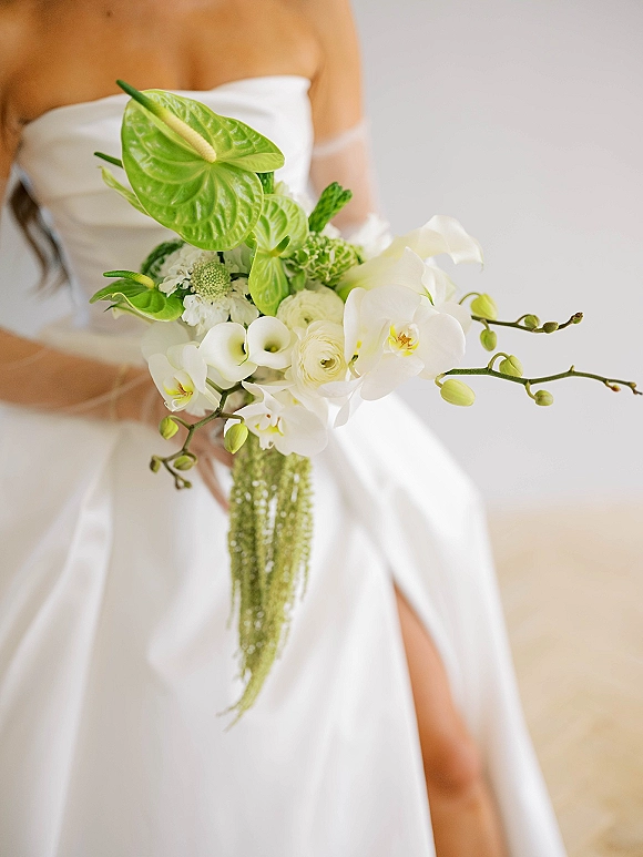 Bridal bouquet of white orchids and calla lilies with cascading amaranthus and greenery held against a strapless dress indoors
