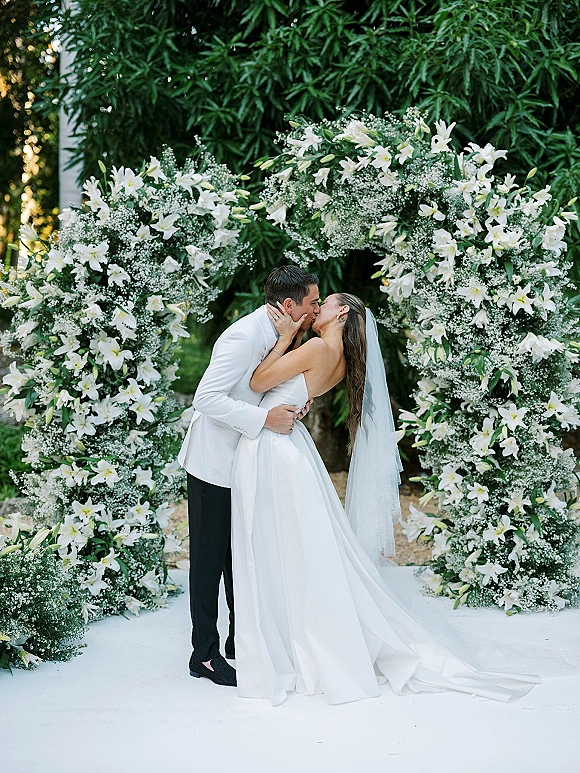 Wedding kiss portrait of bride and groom kiss beneath a crescent arch of white lilies and baby's breath, veil flowing on a garden aisle