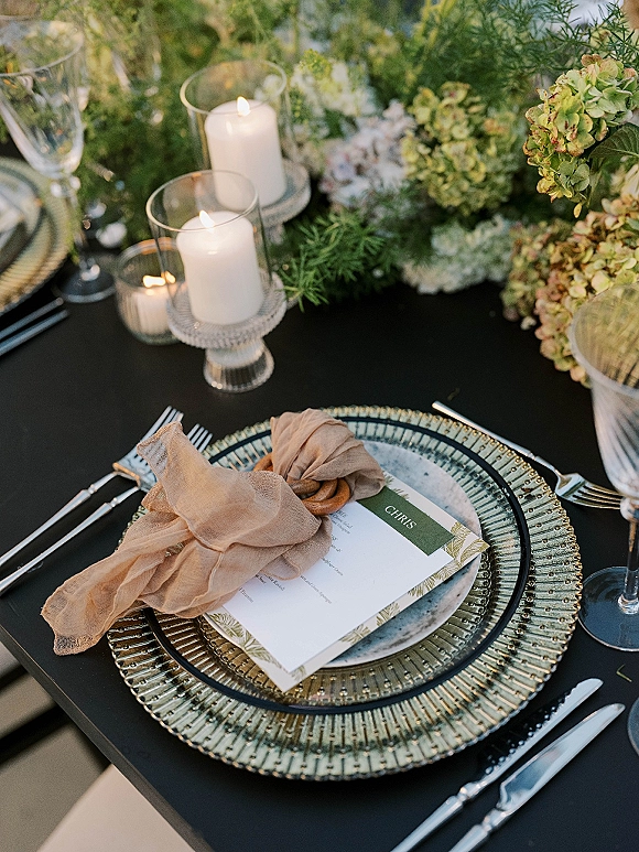 Wedding place setting with black wedding tablescape, menu card on layered plates, napkin bow, candlelight, and floral centerpiece on black linen outdoors