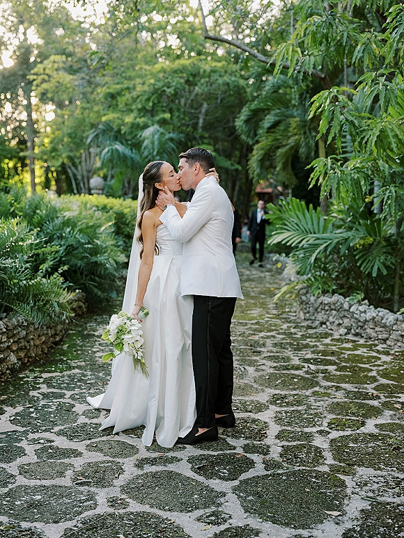 Wedding kiss portrait of bride and groom kissing, bride holding a bouquet with veil accent, on a stone garden path with palm trees