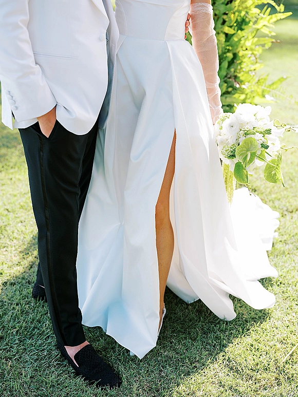 Wedding couple portrait with bride holding a cascading white bouquet in slit dress and sheer gloves beside groom in white tuxedo jacket on sunlit lawn