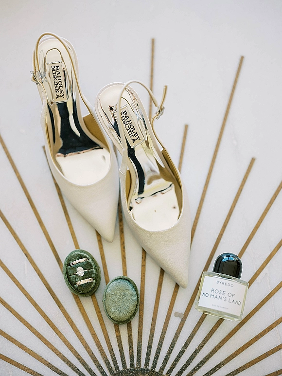 Bridal shoes with metallic insoles in a flat lay beside a velvet ring box, engagement ring, bands and perfume on gold sunburst backdrop