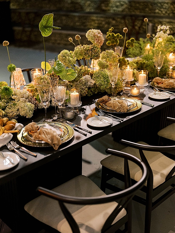 Reception tablescape with a green hydrangea centerpiece, pillar and votive candlelight, gold chargers and crystal glassware on a black table against a stone wall