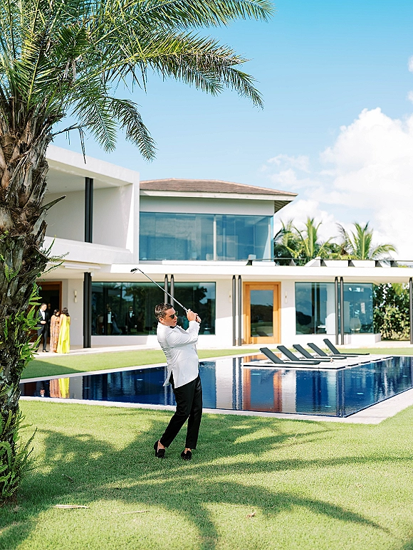 Groom portrait of a man in a white tuxedo and sunglasses holding a golf club by a modern villa pool with palm trees and blue sky