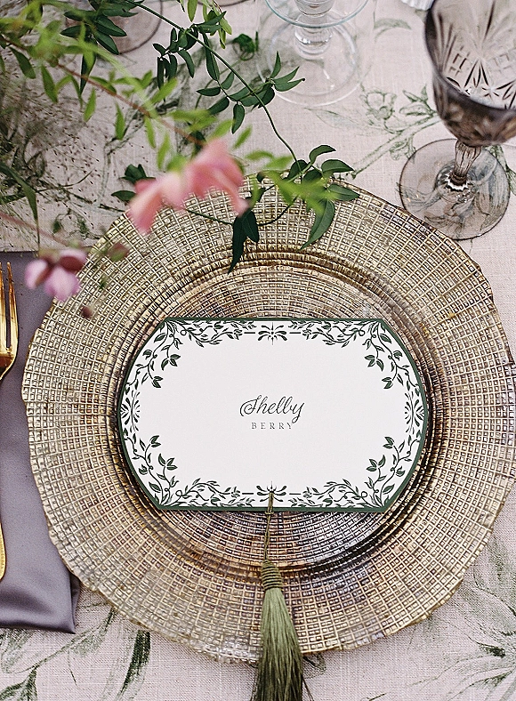 Wedding place setting with a gold charger plate, place card and tassel, gold flatware, and glassware on a floral linen tablecloth