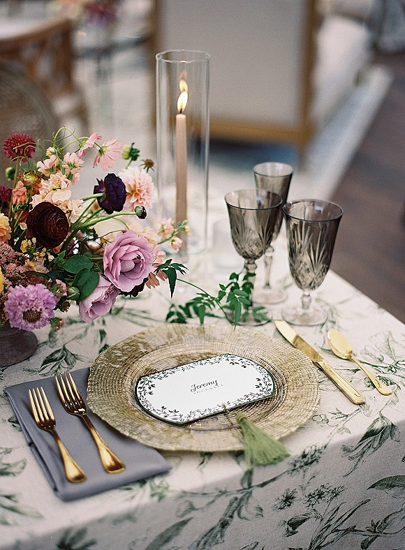 Reception tablescape with wedding place setting, woven charger, gold flatware, gray napkin, smoked goblets, and floral centerpiece candle in glass hurricane