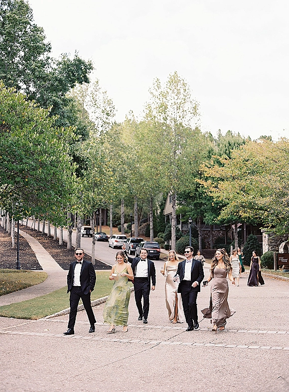 Wedding party portrait of bridesmaids and groomsmen walking in tuxedos and neutral dresses, holding champagne flutes on a tree-lined street