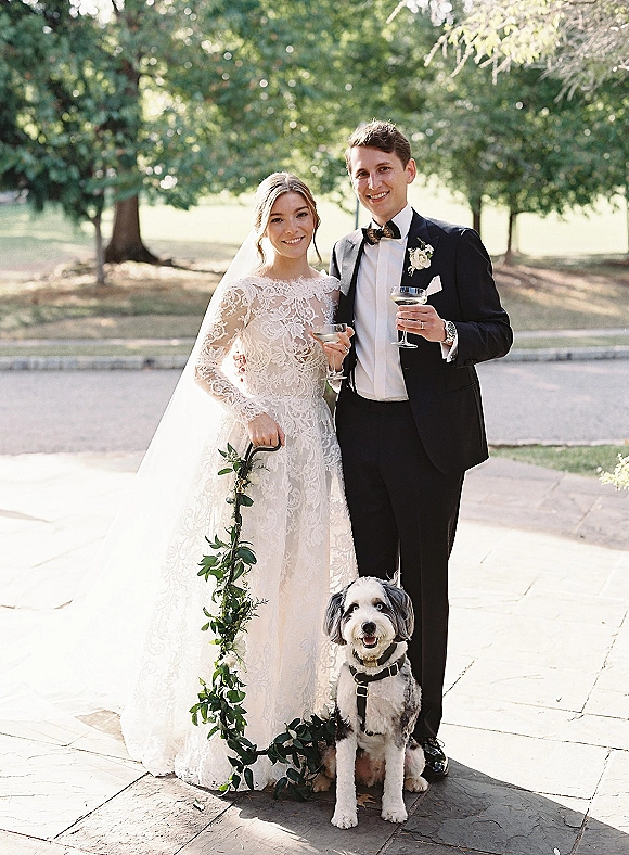 Couple portrait of bride and groom with dog, holding cocktail glasses on a stone patio, bride in lace gown and veil, groom in black tuxedo