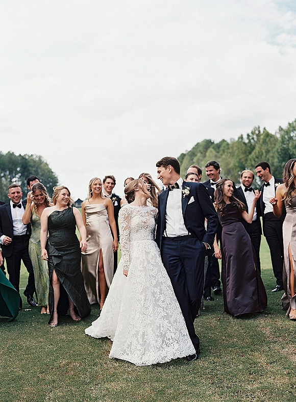 Wedding party portrait of bride and groom with wedding party, bride in long sleeve lace gown, on a grassy field with trees under cloudy sky