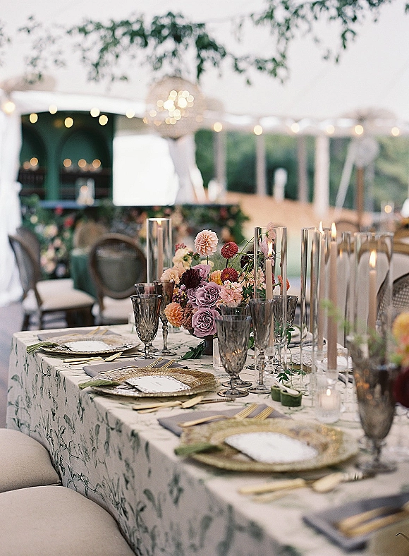 Reception tablescape with wedding table centerpiece, taper candles in glass holders, patterned linens and gold flatware under string-lit tent draping