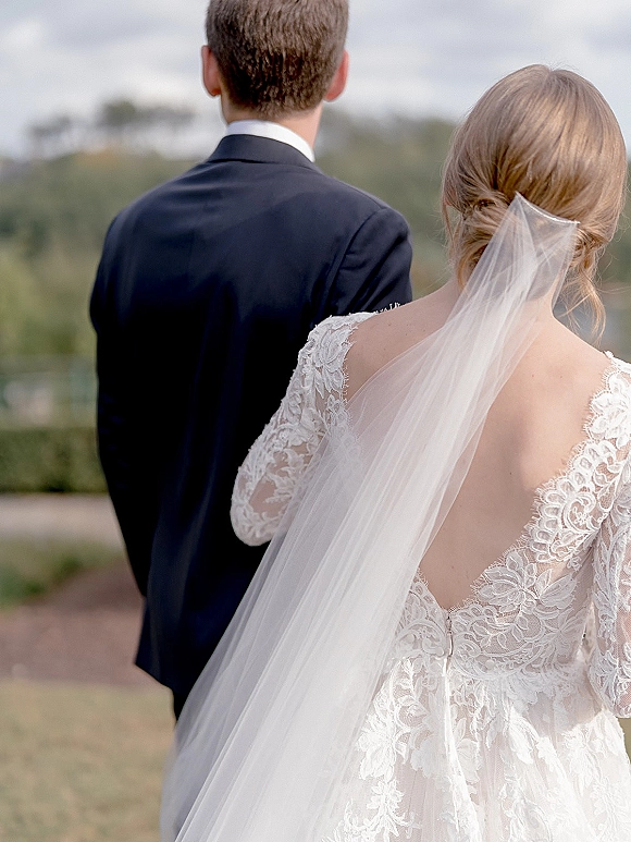 Wedding couple portrait of bride and groom from behind, her cathedral veil over lace dress as they face hills and open sky outdoors