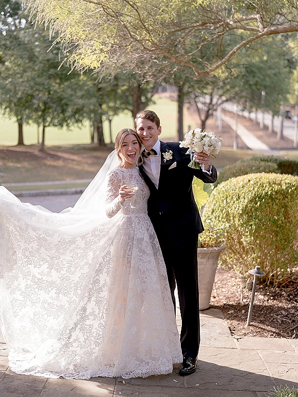 Couple portrait of bride and groom smiling, her long lace veil and bouquet visible as they toast with champagne on a garden patio